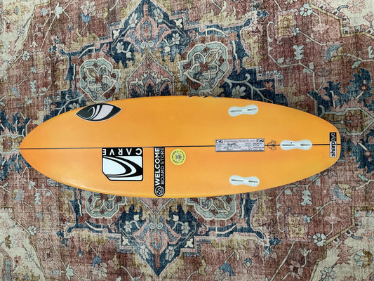Orange Sharpeye HT2 Youth pre-loved surfboard bottom with three fin boxes on a patterned rug