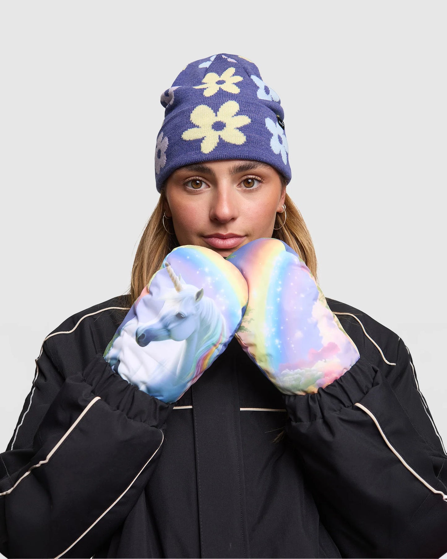 Person wearing a blue floral beanie and colorful mittens with a unicorn design on a plain background