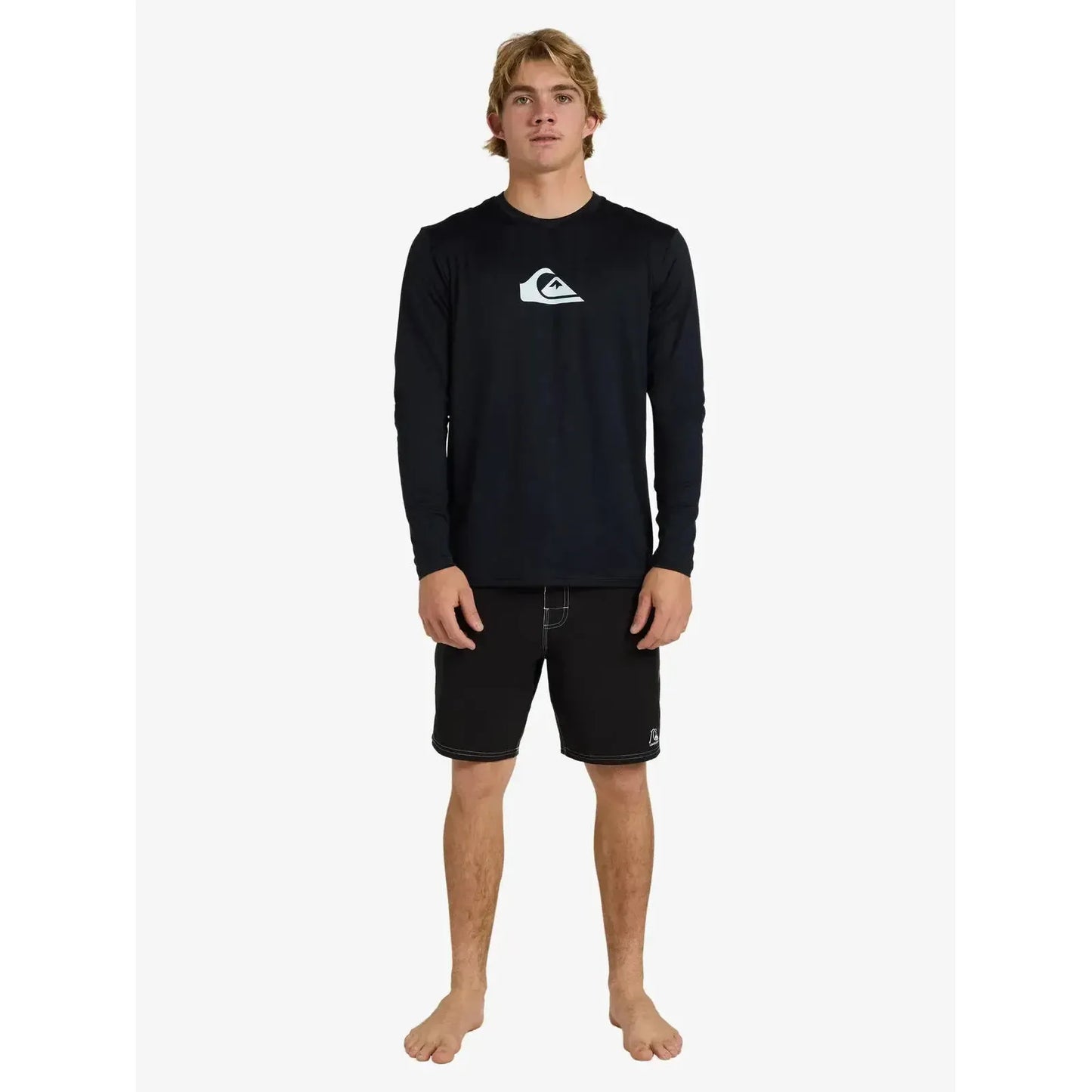 Man wearing long sleeve black surf rash guard and black boardshorts, standing barefoot