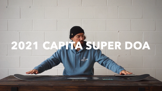 Person in Welcome Snow hoodie reviews 2021 Capita Super DOA snowboard on wooden table