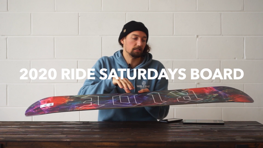Man in blue hoodie displaying 2020 Ride Saturdays snowboard on wooden table indoors