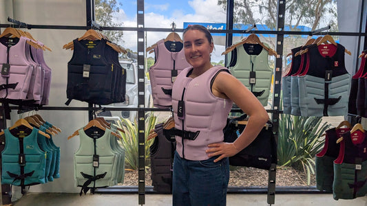 Staff member at Welcome Board Store wearing a snug pink foam life jacket, zipped and buckled, in front of the vest wall