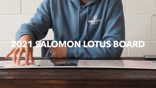 Person in blue Welcome Snow hoodie presenting 2021 Salomon Lotus snowboard on wooden table