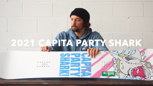 Man in blue hoodie showing 2021 Capita Party Shark snowboard with colorful shark graphic at Welcome Boardstore