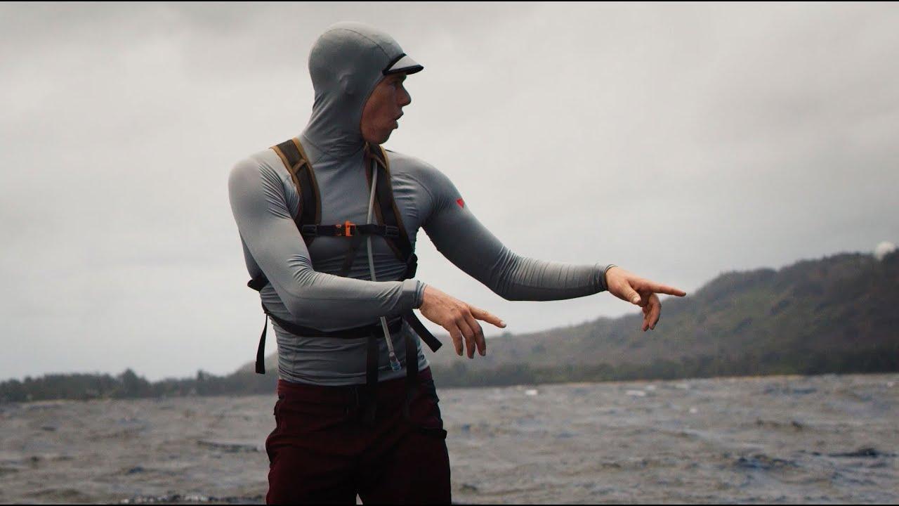 Person in gray hooded surf or wakeboard gear near water, pointing, with cloudy sky and hills.