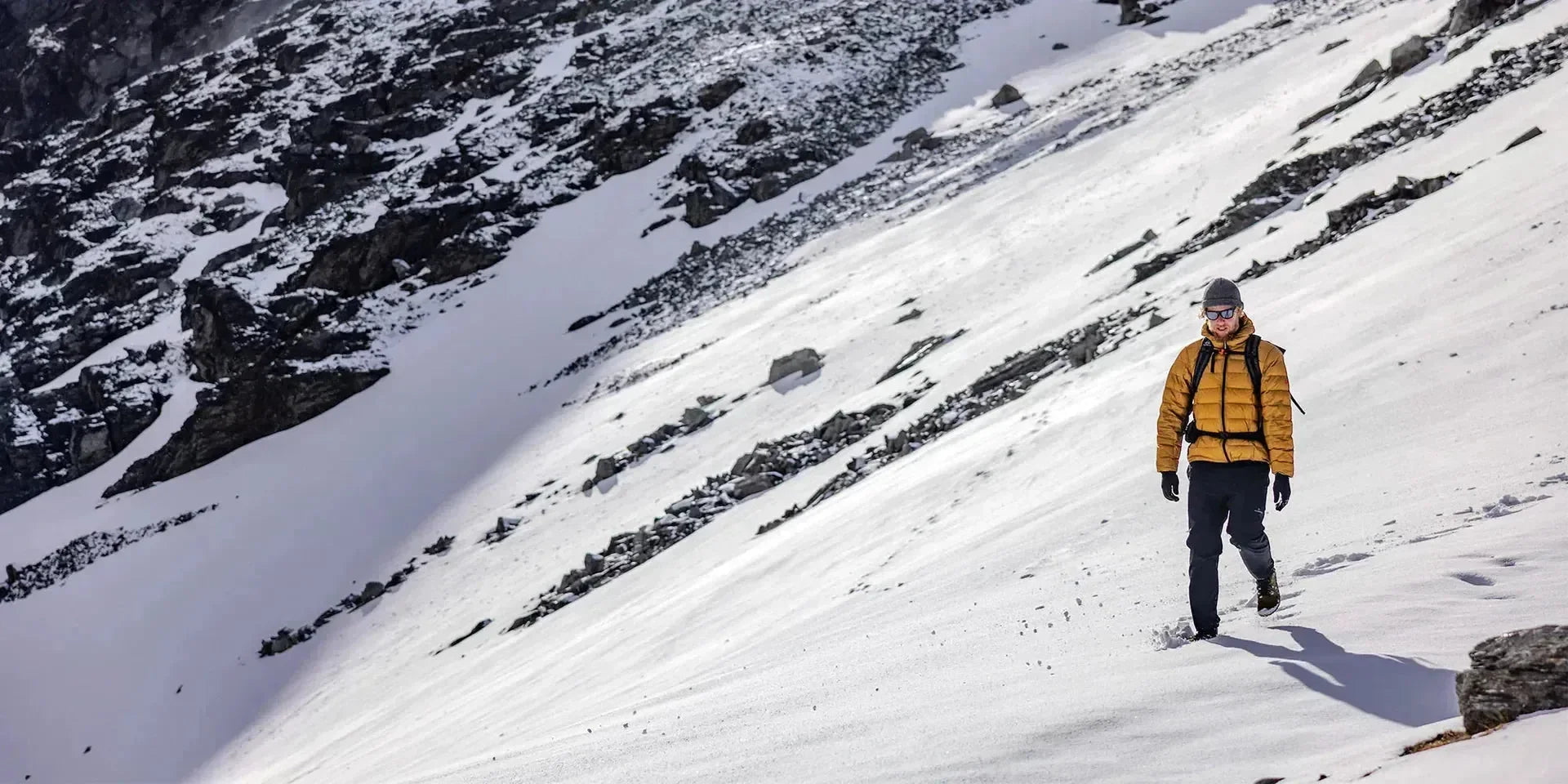 Man hiking in snow wearing yellow insulated jacket, winter pants and sunglasses, snowy mountain slope