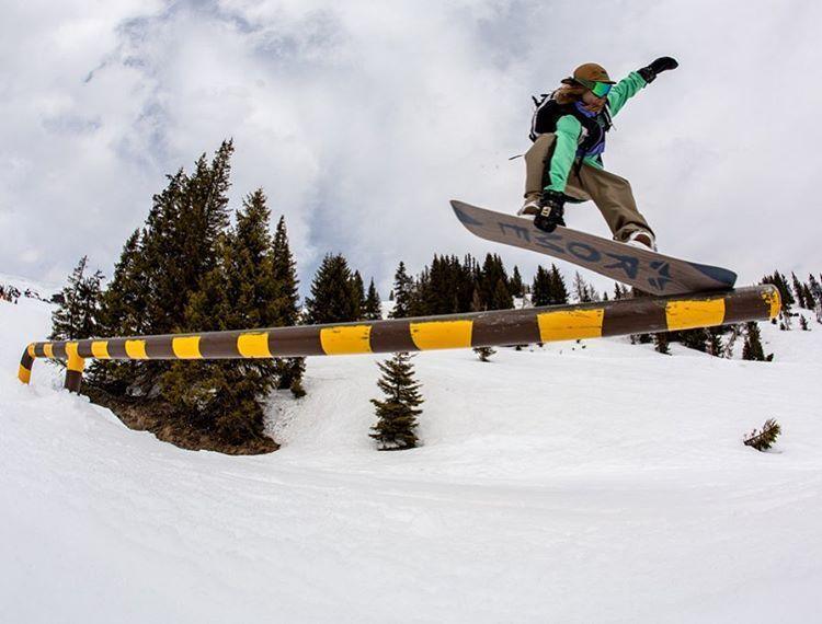 Snowboarder jumping over yellow rail on snowy mountain with trees, snowboarding gear