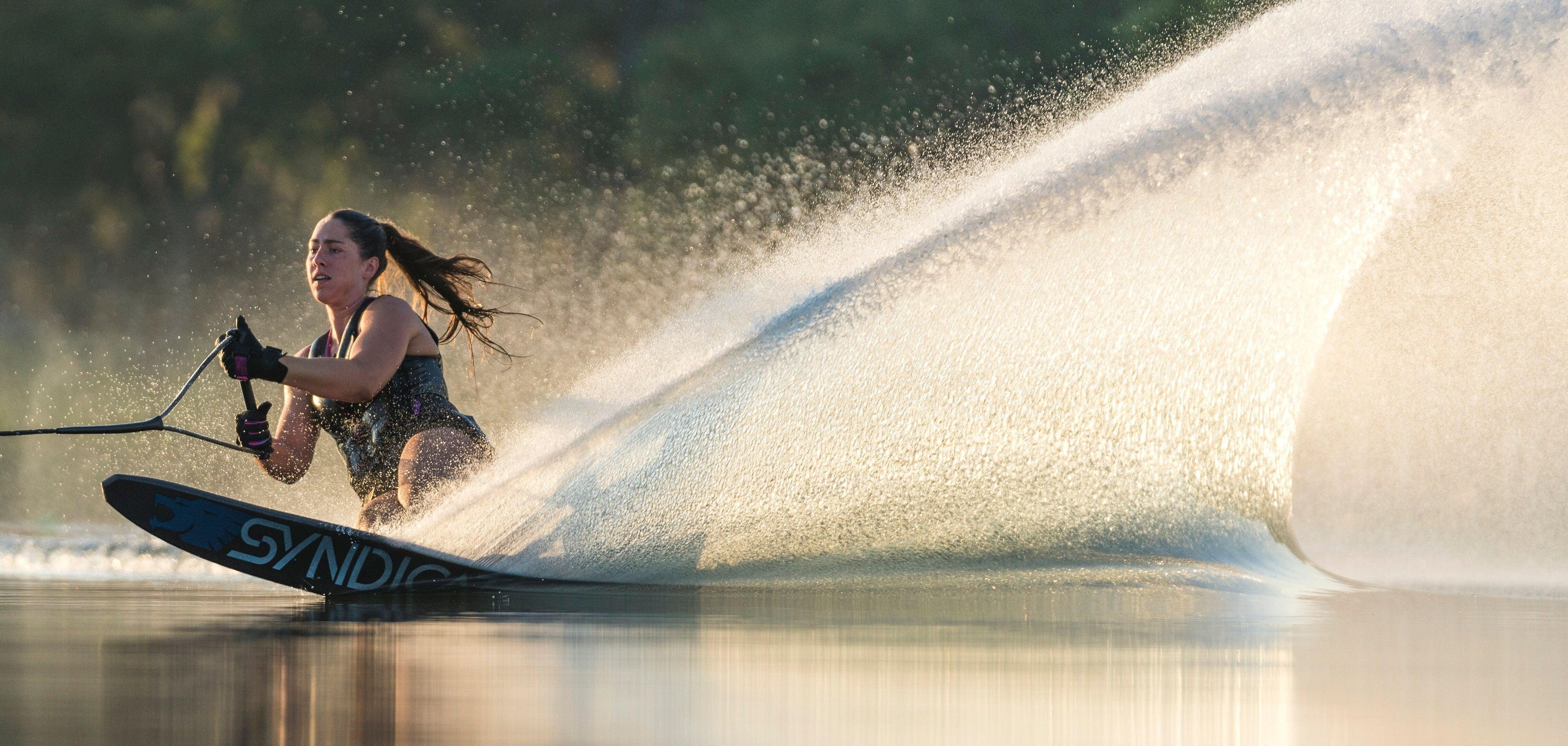 Woman wakeboarding on calm water wearing life vest, creating a spray, wakeboard essentials