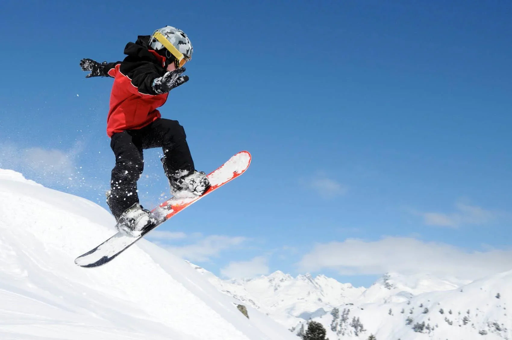 Boy snowboarding in mid-air on snowy mountain, wearing red jacket and helmet, clear sky