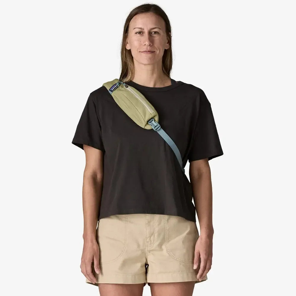 Woman wearing a black t-shirt and beige shorts with a green and blue bag over her shoulder on a white  welcome boardstore background