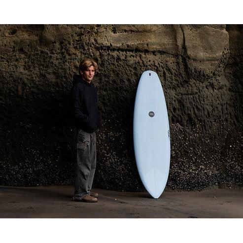 Man in coastal cave standing next to a blue surfboard, surf gear from Welcome Boardstore