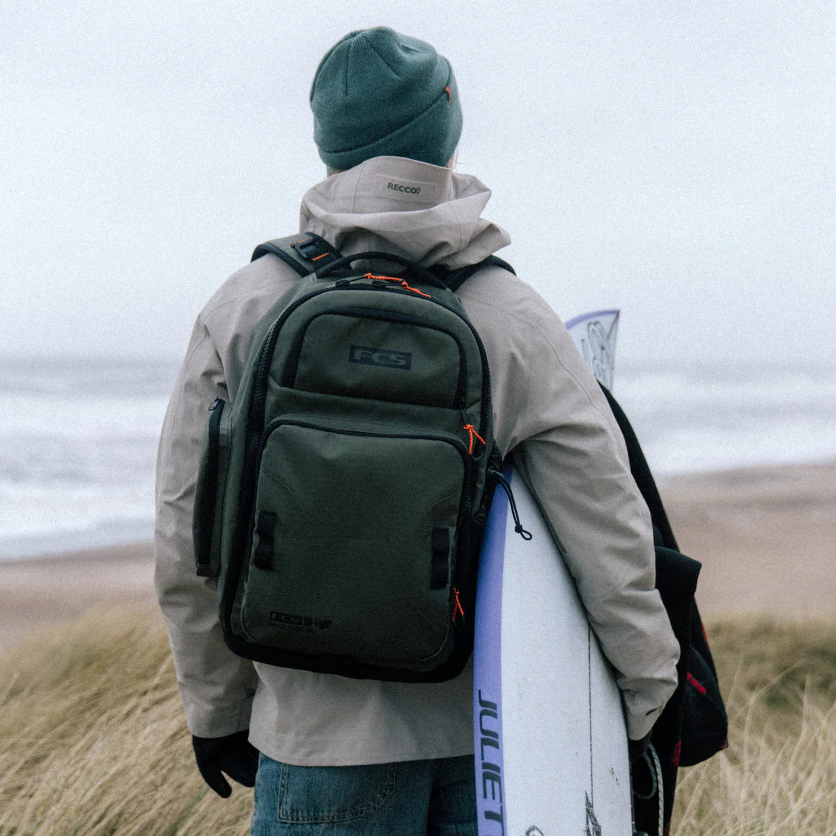 Person with a FCS Day X Pack 24L green backpack and surfboard standing on a beach