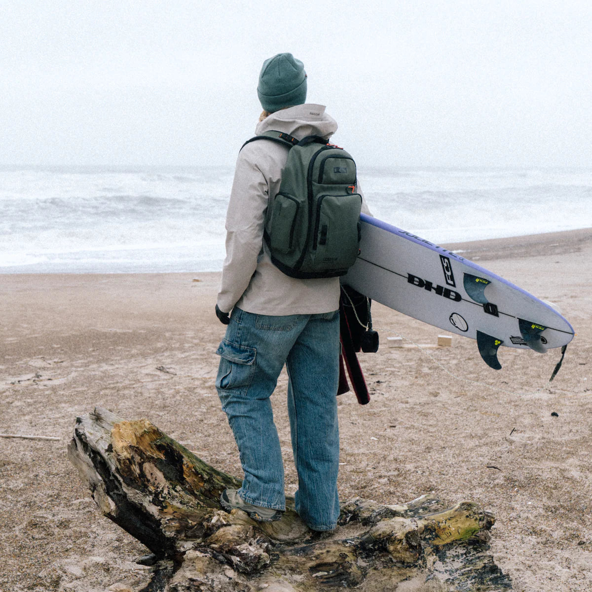 Person holding a surfboard on a beach with a FCS Day X Pack 24L backpack