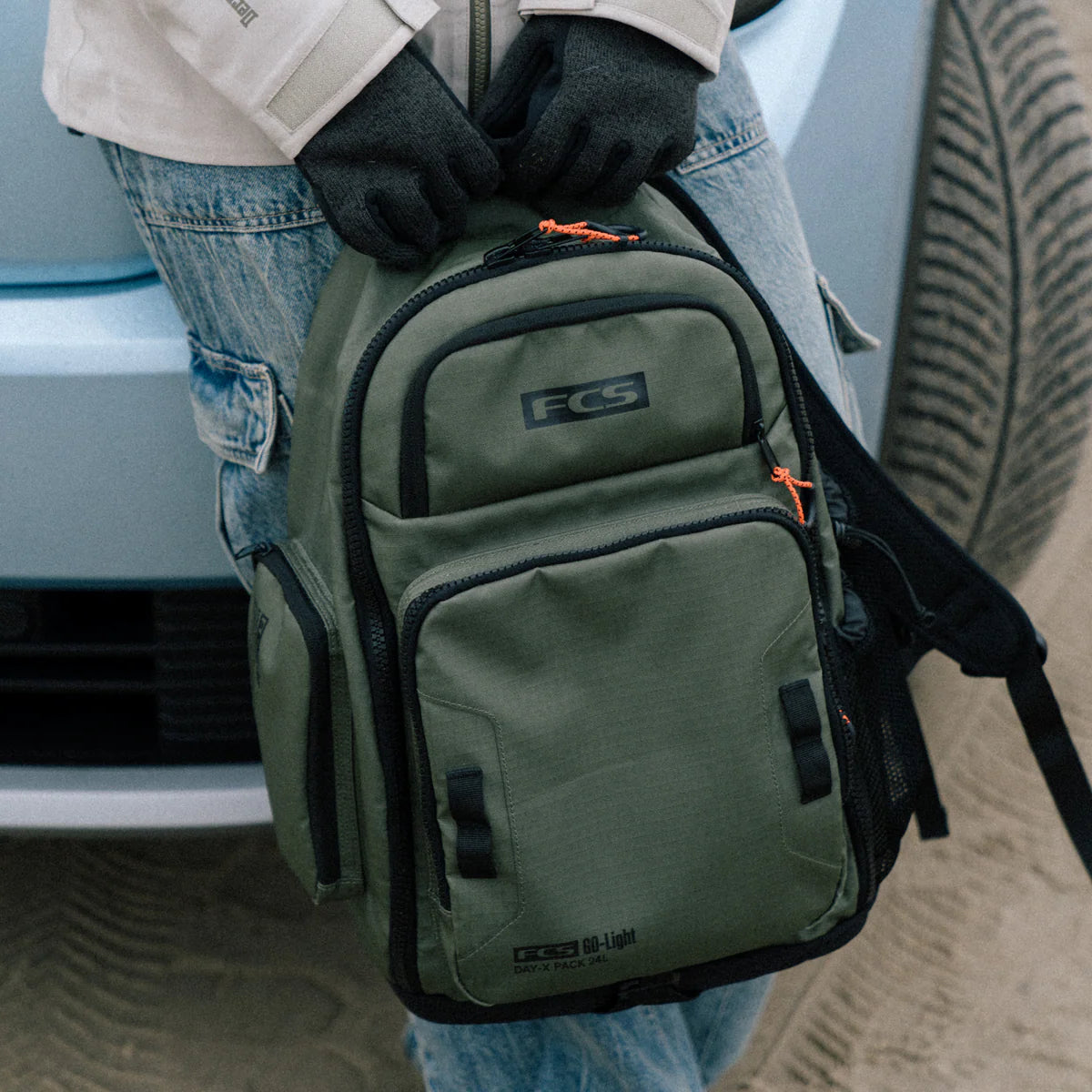 Green backpack with FCS logo held by a person wearing gloves, with a car and sand in the background.