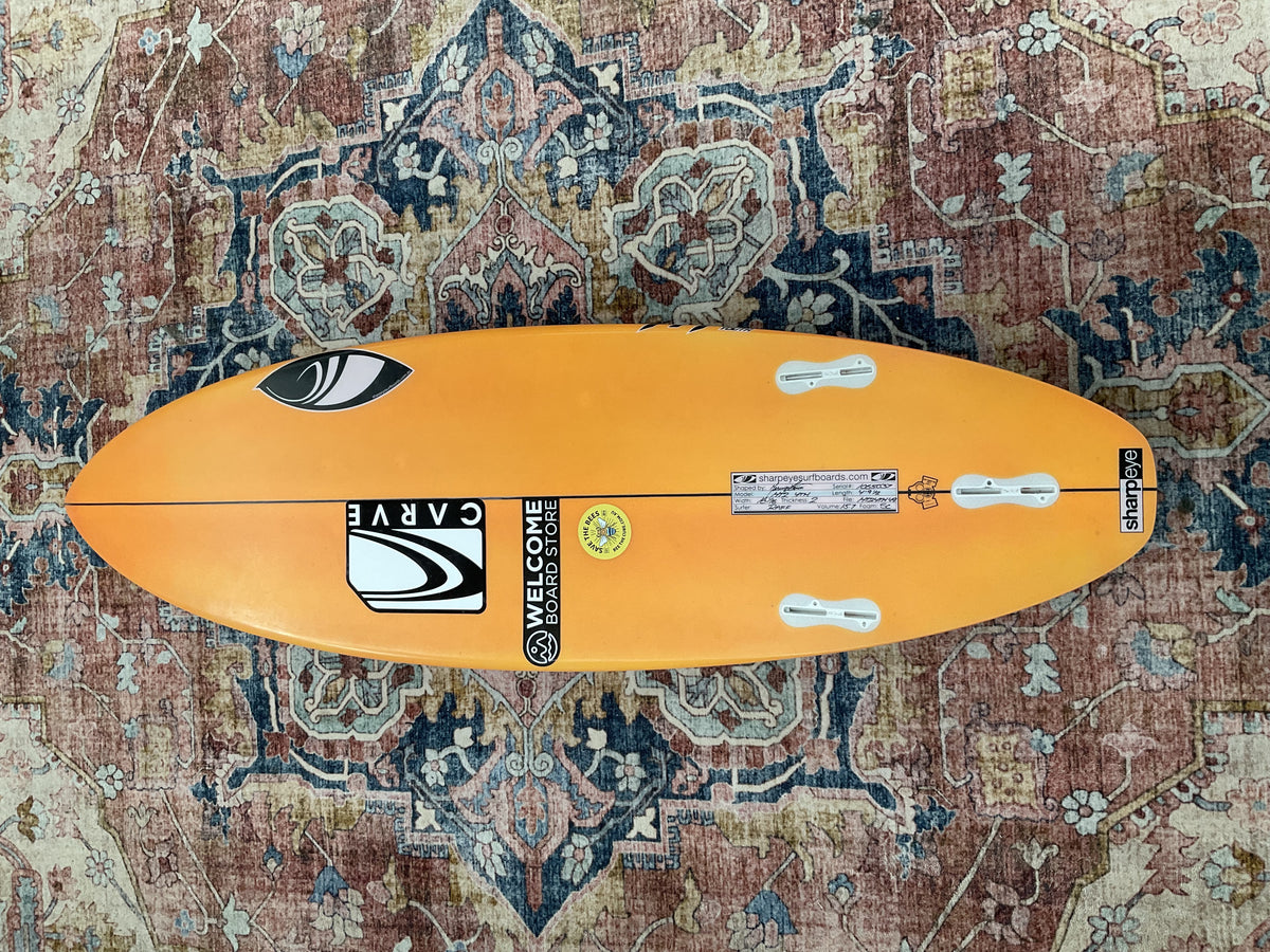 Orange Sharpeye HT2 Youth pre-loved surfboard bottom with three fin boxes on a patterned rug