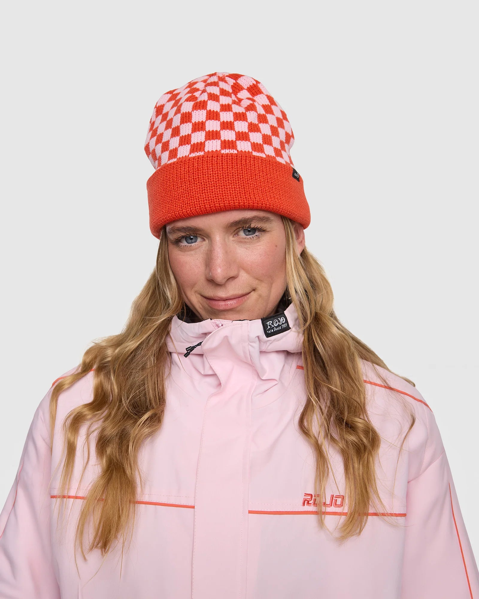 Rojo Cosy Check Beanie Person wearing a red and white checkered beanie and pink jacket with visible branding on a light gray background