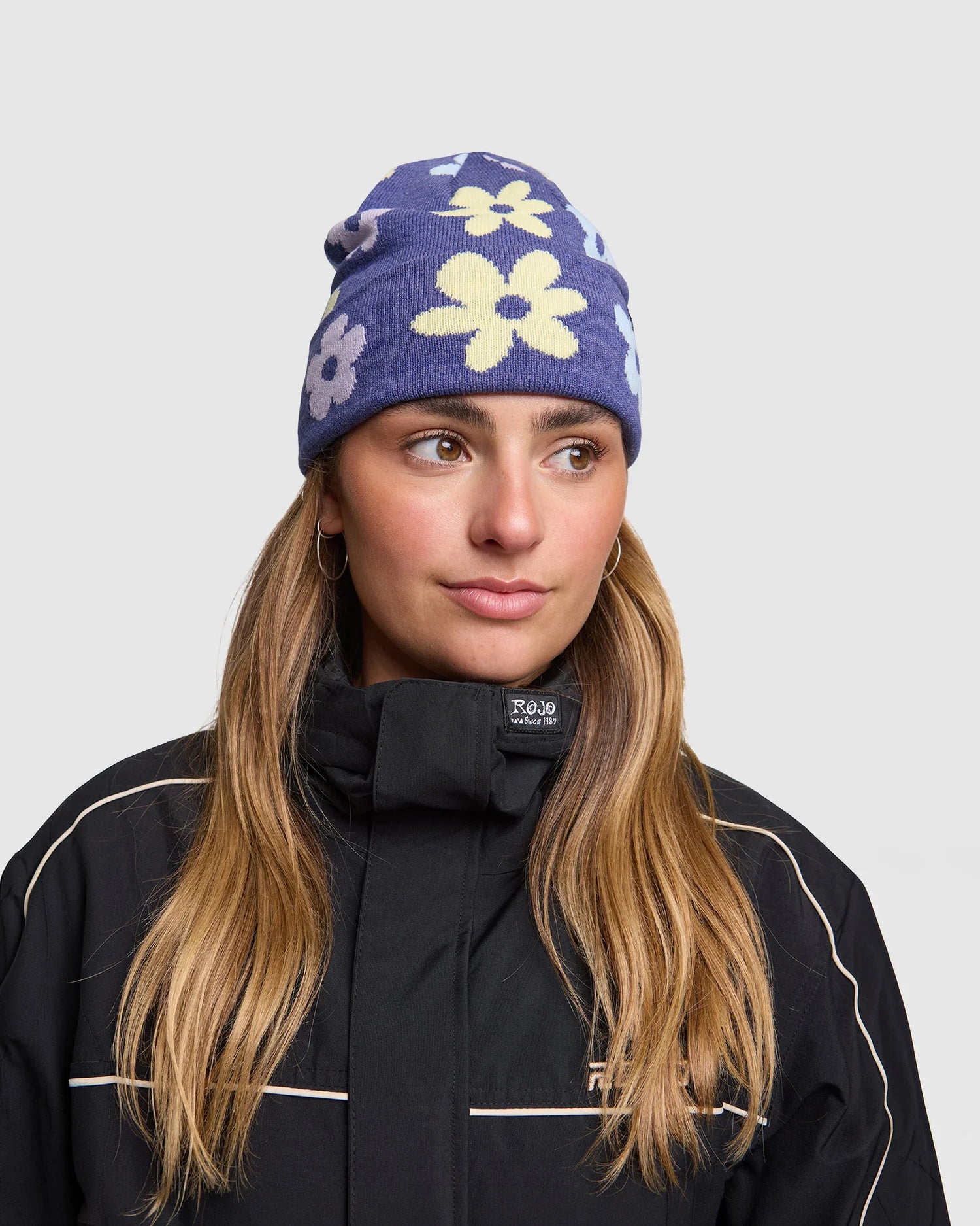 Rojo Spring Bloom Beanie Person wearing a purple beanie with floral patterns and a black jacket on a light gray background